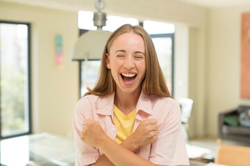 Wall Mural - pretty blond woman smiling cheerfully and celebrating, with fists clenched and arms crossed, feeling happy and positive