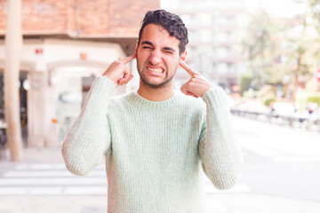 Sticker - young hispanic man looking angry, stressed and annoyed, covering both ears to a deafening noise, sound or loud music