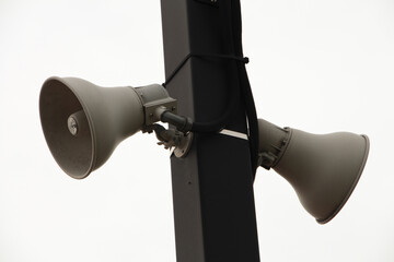 two beige train station speakers pointing in opposite directions fastened to a black rectangular post outside at train station with sky in background