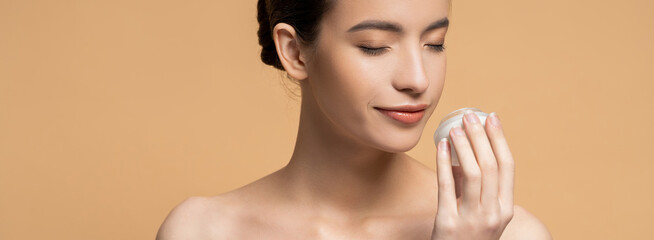 Poster - Young asian woman with naked shoulders smelling cosmetic cream isolated on beige, banner