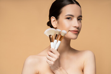 Canvas Print - Brunette asian woman with naked shoulders holding makeup brushes isolated on beige