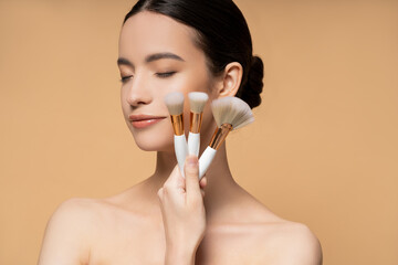 Wall Mural - Young asian woman with closed eyes and naked shoulders holding makeup brushes isolated on beige