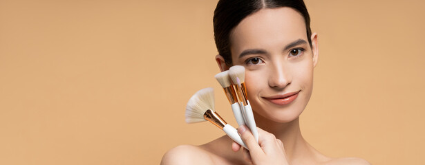 Wall Mural - Smiling asian model with naked shoulders holding makeup brushes isolated on beige, banner