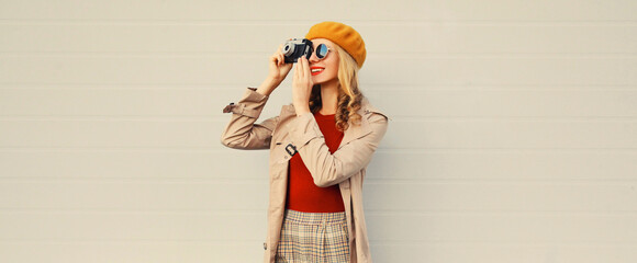 Wall Mural - Fashionable autumn color style outfit, stylish young woman photographer with film camera wearing orange french beret hat, jacket and round sunglasses on gray background