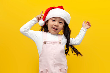 little asian girl in santa hat rejoice and smile on yellow background, korean child holding christmas accessory