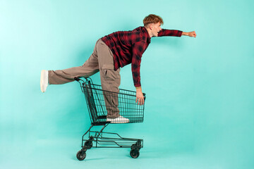 young happy guy shopper sits and rides in shopping cart from supermarket on blue isolated background