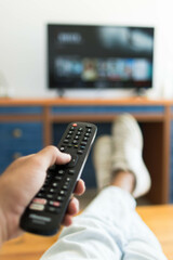 Vertical photo of a hand holding a television remote control while his feet are on the table.