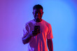 © Богдан Маліцький - african american man is using smartphone and smiling in neon lighting, guy is typing on phone online