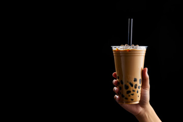 A hand holding takeaway plastic cup of delicious iced bubble milk tea and black pearls isolated on black background with copy space