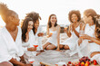 © Prostock-studio - Excited multiracial female friends chatting, enjoying summer picnic with wine or champagne at sunset on beach