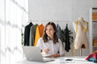 © Natee Meepian - Portrait of tailor woman working on clothes in tailoring atelier. Attractive beautiful young female fashion designer work on laptop in workshop