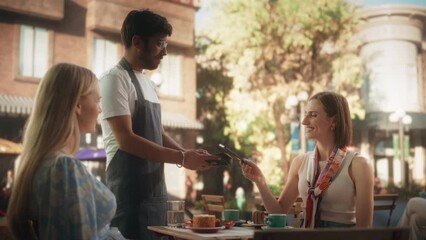 Wall Mural - Two Female Friends Ordering Delicious Cake Desserts From a Member of the Staff in a Cafe. Handsome Multiethnic Waiter Holding a Terminal, Female Using Her Smartphone for a Contactless NFC Payment