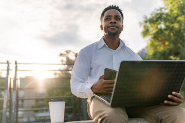 After a tiring day at work, a young businessman sits by the riverside, engrossed in his mobile phone and laptop, checking the social media platforms he frequents.