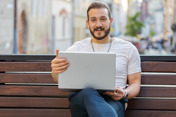 Portrait of freelancer man opens laptop sitting on bench in urban city street outdoors. Middle eastern guy works on notebook, sends messages, makes online purchases, watching movies. Town lifestyles