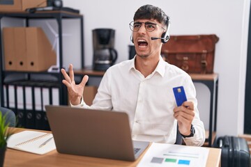 Wall Mural - Young hispanic man working using computer laptop holding credit card crazy and mad shouting and yelling with aggressive expression and arms raised. frustration concept.