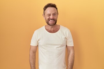 Wall Mural - Middle age man with beard standing over yellow background winking looking at the camera with sexy expression, cheerful and happy face.