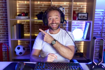 Poster - Middle age man with beard playing video games wearing headphones cheerful with a smile on face pointing with hand and finger up to the side with happy and natural expression