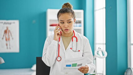 Canvas Print - Young beautiful hispanic woman doctor talking on smartphone with relaxed expression reading medical report at clinic