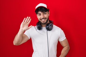 Poster - Hispanic man with beard wearing gamer hat and headphones waiving saying hello happy and smiling, friendly welcome gesture