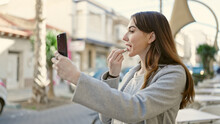 Young Hispanic Woman Using Smartphone As A Mirror Applying Lipstick At Coffee Shop Terrace