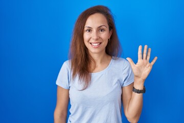 Canvas Print - Brunette woman standing over blue background showing and pointing up with fingers number five while smiling confident and happy.