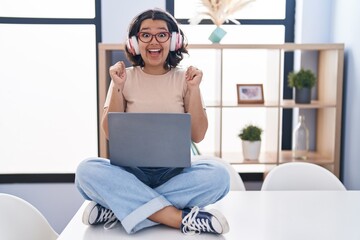 Poster - Young hispanic woman using laptop sitting on the table wearing headphones celebrating surprised and amazed for success with arms raised and open eyes. winner concept.