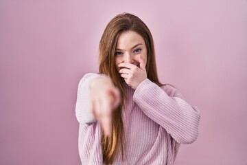 Wall Mural - Young caucasian woman standing over pink background laughing at you, pointing finger to the camera with hand over mouth, shame expression