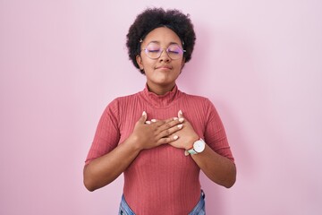 Sticker - Beautiful african woman with curly hair standing over pink background smiling with hands on chest with closed eyes and grateful gesture on face. health concept.