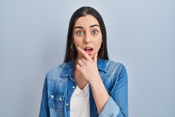 Poster - Hispanic woman standing over blue background looking fascinated with disbelief, surprise and amazed expression with hands on chin