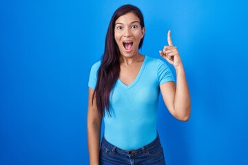 Sticker - Young hispanic woman standing over blue background pointing finger up with successful idea. exited and happy. number one.