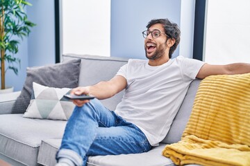 Sticker - Handsome latin man holding television remote control celebrating crazy and amazed for success with open eyes screaming excited.