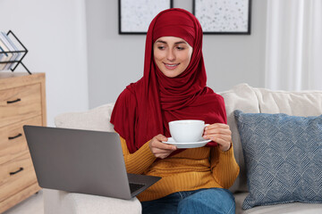 Wall Mural - Muslim woman with cup of drink using laptop at couch in room