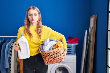 Poster - Young caucasian woman holding laundry basket and detergent bottle relaxed with serious expression on face. simple and natural looking at the camera.