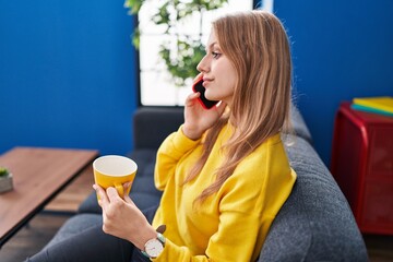 Wall Mural - Young blonde woman talking on smartphone drinking coffee at home