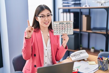 Sticker - Chinese young woman working at the office holding holidays calendar surprised with an idea or question pointing finger with happy face, number one