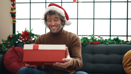 Sticker - Young hispanic man looking gift sitting on sofa by christmas decoration at home