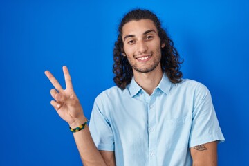 Poster - Young hispanic man standing over blue background smiling with happy face winking at the camera doing victory sign with fingers. number two.