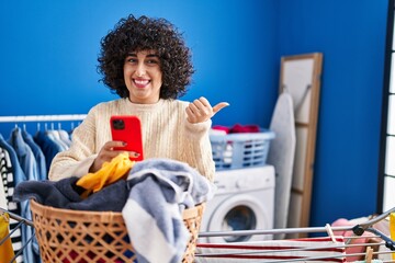 Wall Mural - Young brunette woman with curly hair doing laundry using smartphone pointing thumb up to the side smiling happy with open mouth