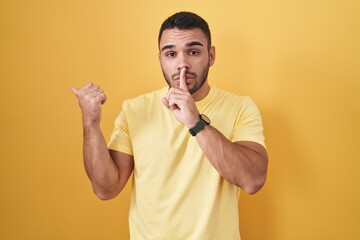 Poster - Young hispanic man standing over yellow background asking to be quiet with finger on lips pointing with hand to the side. silence and secret concept.