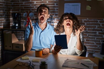 Poster - Middle age hispanic couple using touchpad sitting on the table at night crazy and mad shouting and yelling with aggressive expression and arms raised. frustration concept.