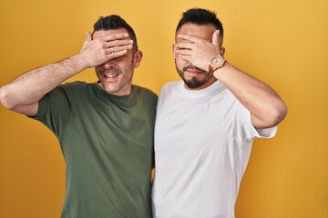 Canvas Print - Homosexual couple standing over yellow background smiling and laughing with hand on face covering eyes for surprise. blind concept.