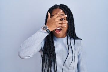 Wall Mural - African american woman standing over blue background peeking in shock covering face and eyes with hand, looking through fingers with embarrassed expression.