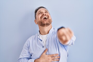 Sticker - Middle age caucasian man standing over blue background laughing at you, pointing finger to the camera with hand over body, shame expression