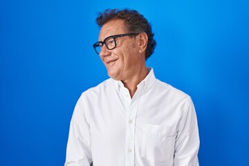 Wall Mural - Middle age hispanic man standing over blue background looking away to side with smile on face, natural expression. laughing confident.