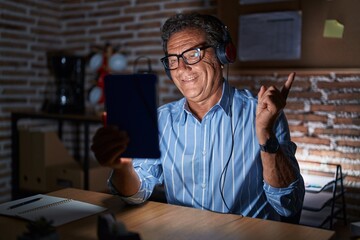 Sticker - Middle age hispanic man using touchpad sitting on the table at night with a big smile on face, pointing with hand finger to the side looking at the camera.