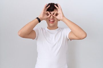 Poster - Young non binary man wearing casual white t shirt doing ok gesture like binoculars sticking tongue out, eyes looking through fingers. crazy expression.