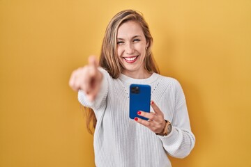 Wall Mural - Young blonde woman using smartphone typing message pointing to you and the camera with fingers, smiling positive and cheerful