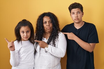 Sticker - Family of mother, daughter and son standing over yellow background pointing with hand finger to the side showing advertisement, serious and calm face