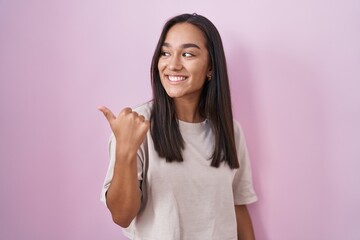 Sticker - Young hispanic woman standing over pink background smiling with happy face looking and pointing to the side with thumb up.