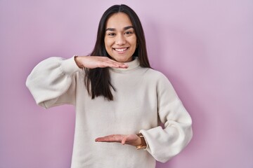 Wall Mural - Young south asian woman standing over pink background gesturing with hands showing big and large size sign, measure symbol. smiling looking at the camera. measuring concept.